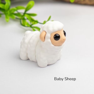 Puede incluir: Figura de oveja blanca hecha a mano con cuerpo esponjoso y cara, orejas y ojos negros grandes de color beige. El texto "Baby Sheep" est&aacute; impreso debajo.
