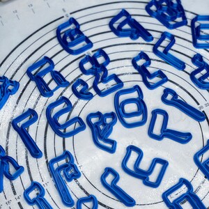May include: Blue plastic cookie cutters shaped like letters of the Hebrew alphabet on a white surface with flour dusted on it.