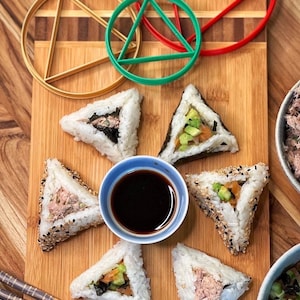 May include: Six triangular sushi rice balls on a wooden cutting board with soy sauce and chopsticks. The sushi is filled with avocado, cucumber, and tuna. There are three plastic geometric shapes in the background: a red triangle, a green circle, and a gold circle.