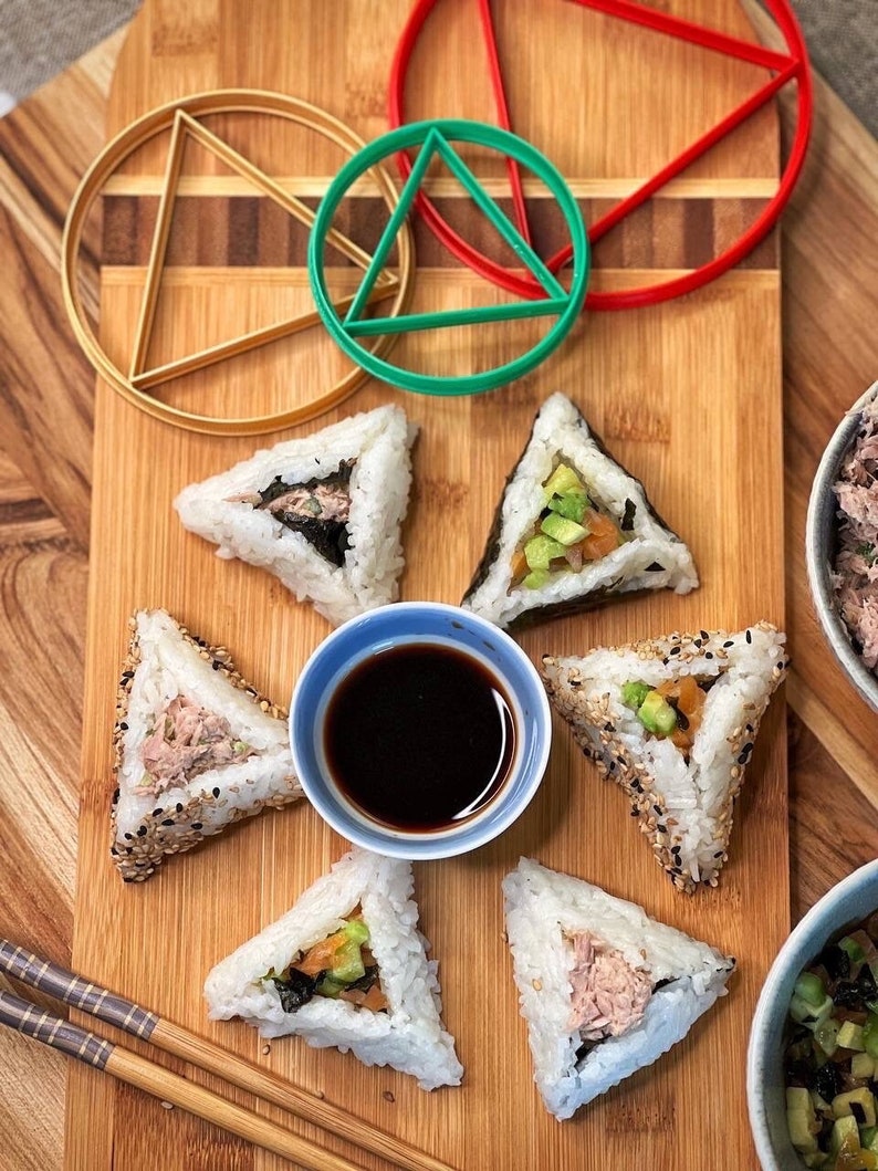 May include: Six triangular sushi rice balls on a wooden cutting board with soy sauce and chopsticks. The sushi is filled with avocado, cucumber, and tuna. There are three plastic geometric shapes in the background: a red triangle, a green circle, and a gold circle.