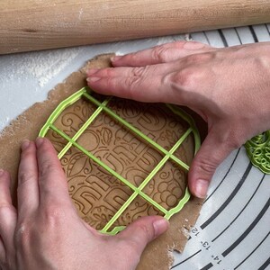 May include: A person using a green plastic cookie cutter to cut out a gingerbread cookie. The cookie cutter has a grid pattern and the words "Happy" and "Holidays" are visible on the cookie dough.