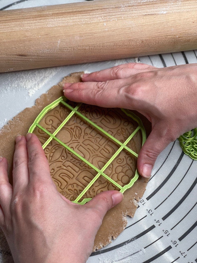 May include: A person using a green plastic cookie cutter to cut out a gingerbread cookie. The cookie cutter has a grid pattern and the words "Happy" and "Holidays" are visible on the cookie dough.