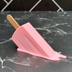 Puede incluir: Un helado de paleta rosa con un palo de madera, derriti&eacute;ndose sobre una superficie de m&aacute;rmol negro.