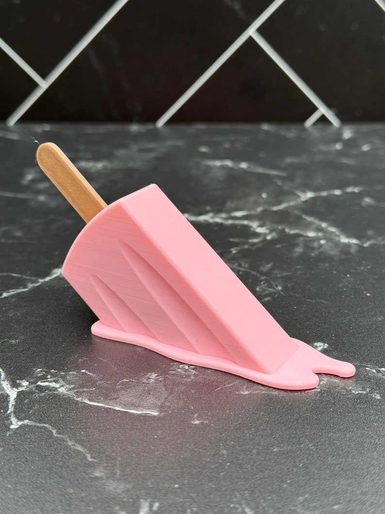 Puede incluir: Un helado de paleta rosa con un palo de madera, derriti&eacute;ndose sobre una superficie de m&aacute;rmol negro.