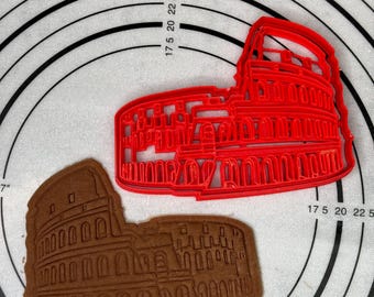Könnte beinhalten: Ein roter Plastik-Ausstecher in Form des Kolosseums in Rom, daneben ein brauner Lebkuchen in Form des Kolosseums auf einer weißen Backmatte mit schwarzen konzentrischen Kreisen.