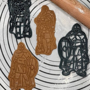 May include: Three cookie cutters in the shape of Viking warriors. The cutters are made of black plastic and are on a white surface with a rolling pin and flour.