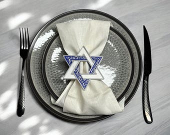 May include: A white napkin with a blue and white Star of David napkin ring on a grey textured plate. The plate is on a white surface.