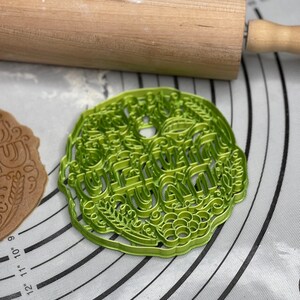 May include: Green plastic cookie cutter with the word "Grandma" in a decorative font. The cookie cutter is on a grey and white silicone baking mat.