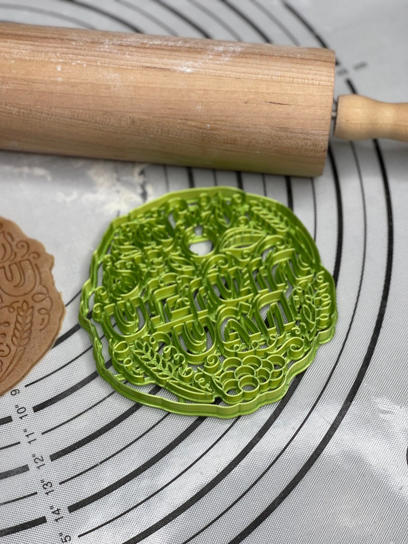 May include: Green plastic cookie cutter with the word "Grandma" in a decorative font. The cookie cutter is on a grey and white silicone baking mat.