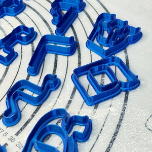 May include: A set of blue plastic cookie cutters in various shapes, including letters, numbers, and abstract designs. The cutters are on a white surface with a grid pattern.