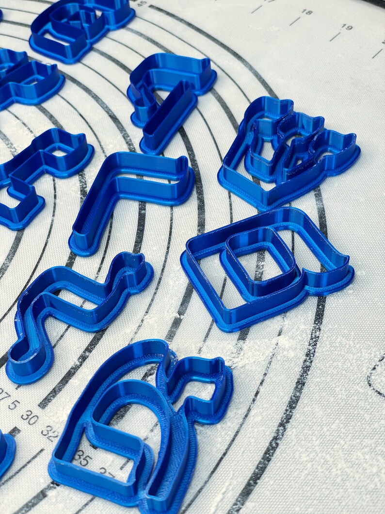 May include: A set of blue plastic cookie cutters in various shapes, including letters, numbers, and abstract designs. The cutters are on a white surface with a grid pattern.