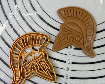 Könnte beinhalten: Zwei goldene Metall-Ausstecher in Form eines spartanischen Helms. Die Ausstecher befinden sich auf einer weißen Silikon-Backmatte mit schwarzen konzentrischen Kreisen.