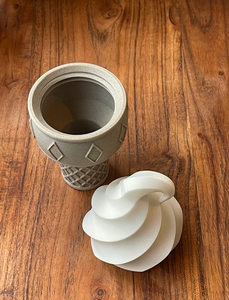 Op de afbeelding: Een witte keramische ijshoornvormige container met een afneembaar deksel. Het deksel is gevormd als een swirl van ijs.