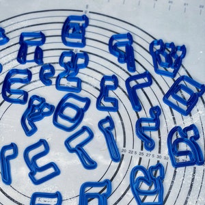 May include: A set of blue plastic cookie cutters shaped like Hebrew letters. The cutters are scattered on a white surface with a circular pattern.