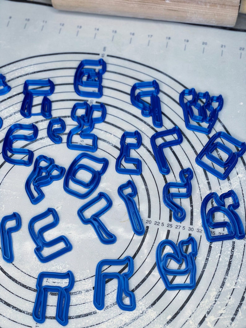 May include: A set of blue plastic cookie cutters shaped like Hebrew letters. The cutters are scattered on a white surface with a circular pattern.