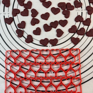 May include: A red heart-shaped cookie cutter set on a white silicone baking mat with a grid pattern. The cookie cutter is made of red plastic and has a grid pattern. There are 42 heart-shaped cookie cutters in the set.