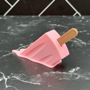 Puede incluir: Un helado de paleta rosa con un palo de madera, derriti&eacute;ndose sobre una superficie de m&aacute;rmol blanco y negro.