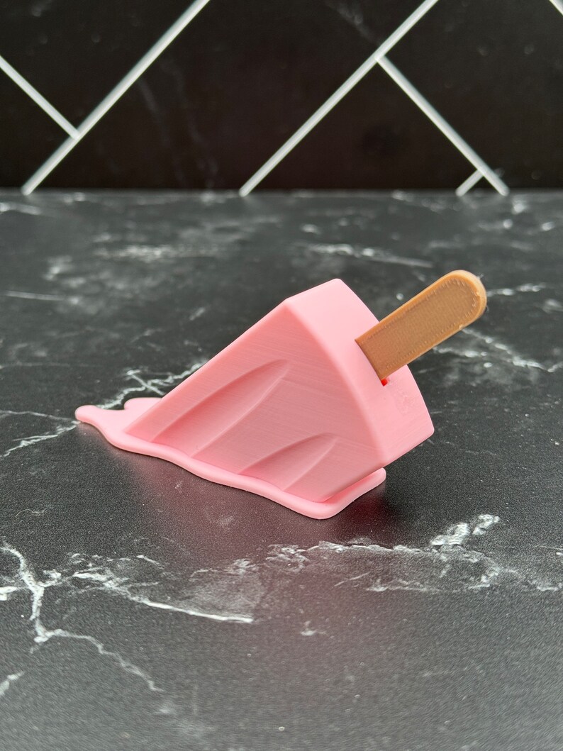 Puede incluir: Un helado de paleta rosa con un palo de madera, derriti&eacute;ndose sobre una superficie de m&aacute;rmol blanco y negro.