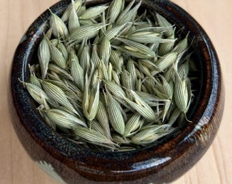 May include: A small, round, dark brown ceramic bowl with a glossy finish filled with a collection of light green, elongated oat grains. The grains are pointed at both ends and feature subtle, vertical, lighter-colored stripes along their surface.