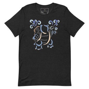 Puede incluir: Camiseta gris oscuro con una ilustración estilizada de una criatura con acentos azules, beige y marrón. El diseño incluye elementos arremolinados similares al agua. La camiseta tiene cuello redondo y mangas cortas.