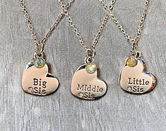 May include: Three silver necklaces with heart-shaped pendants. Each pendant has a small, colourful gem and the words "Big Sis", "Middle Sis", and "Little Sis" engraved on them. The necklaces are displayed on a grey surface.