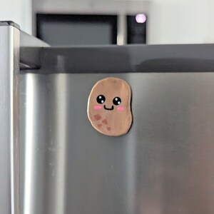 May include: A brown potato-shaped magnet with a painted face, smiling with pink cheeks and black eyes. The magnet is attached to a stainless steel refrigerator.