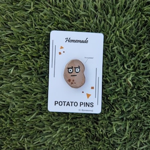 May include: A white card with the text "Homemade Potato Pins" and the text "I'm a potato" above a brown potato pin with a face drawn on it. The card has a brown and orange design on the top and bottom.