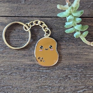 Può includere: Un portachiavi dorato con un ciondolo a forma di patata marrone. La patata ha un viso da cartone animato carino con occhi neri, un blush rosa e un piccolo sorriso.