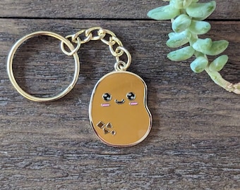 May include: A gold-colored keychain with a brown potato charm. The potato has a cute cartoon face with black eyes, a pink blush, and a small smile.