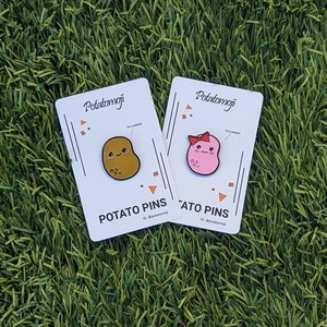 May include: Two potato-shaped enamel pins on white cards, set against a green background. One pin is brown with a smiling face, the other is pink with a bow. The cards read "Potatomoji" and "Potato Pins."