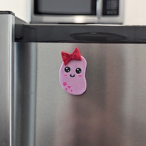 May include: A pink bean-shaped figurine with a red bow, smiling with closed eyes. The figurine has pink and white details and is attached to a silver surface.