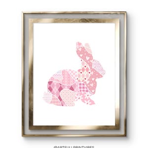 Op de afbeelding: Een roze patchwork konijnen silhouet in een gouden lijst. Het konijn is gemaakt van verschillende roze en witte patronen, waaronder harten, stippen en ruitjes.