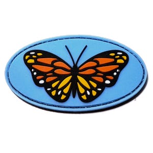 May include: An oval-shaped patch featuring a monarch butterfly graphic with orange, yellow, and black wings. The butterfly is centered on a solid light blue background with a dark blue border, creating a distinct contrast in colors and shapes.