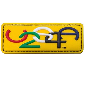 Pode incluir: Um remendo retangular amarelo com as letras "U24A" em um design de elo de corrente. As letras são de diferentes cores: vermelho, verde, branco, preto e azul. O remendo tem uma borda costurada e o símbolo de marca registrada "TM" no canto inferior direito.