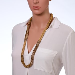 May include: A long, gold-toned necklace with a braided design. The necklace is composed of small, round beads and hangs down the front of a white collared shirt. The necklace is a statement piece.