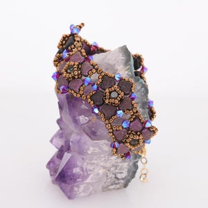 May include: A beaded bracelet with a geometric design featuring purple, brown, and iridescent blue beads. The bracelet is displayed on a purple amethyst crystal.
