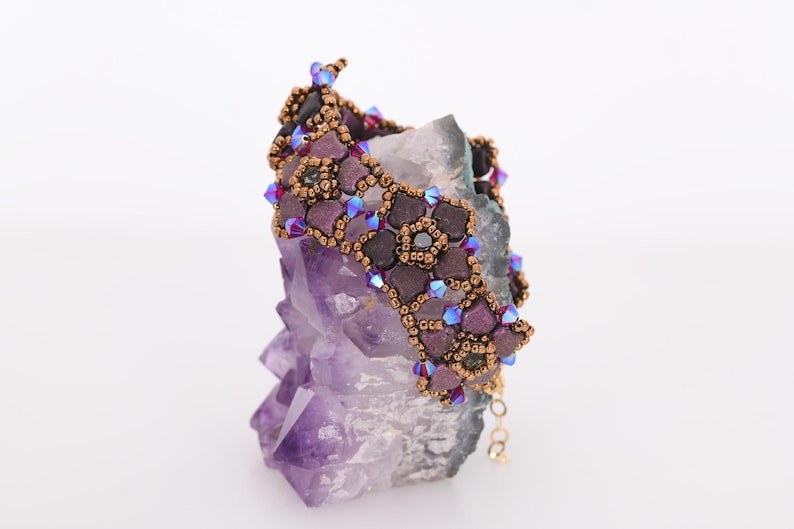 May include: A beaded bracelet with a geometric design featuring purple, brown, and iridescent blue beads. The bracelet is displayed on a purple amethyst crystal.