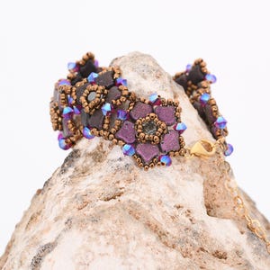 May include: A beaded bracelet with a floral design. The bracelet is made with brown, purple, and blue beads. The bracelet is on a rock.