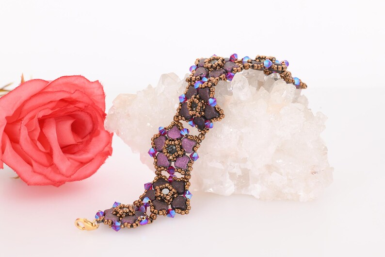 May include: A purple and brown beaded bracelet with a flower design. The bracelet is on a white background with a pink rose.