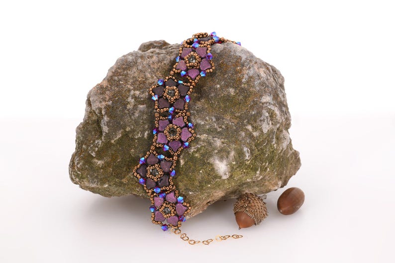 May include: A purple and brown beaded necklace with a floral design. The necklace is on a rock with acorns.