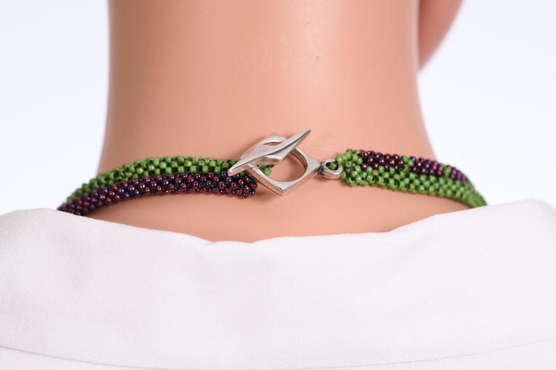 K&ouml;nnte beinhalten: Nahaufnahme eines silbernen Verschlusses an einer Perlenkette. Die Kette besteht aus gr&uuml;nen und violetten Perlen.