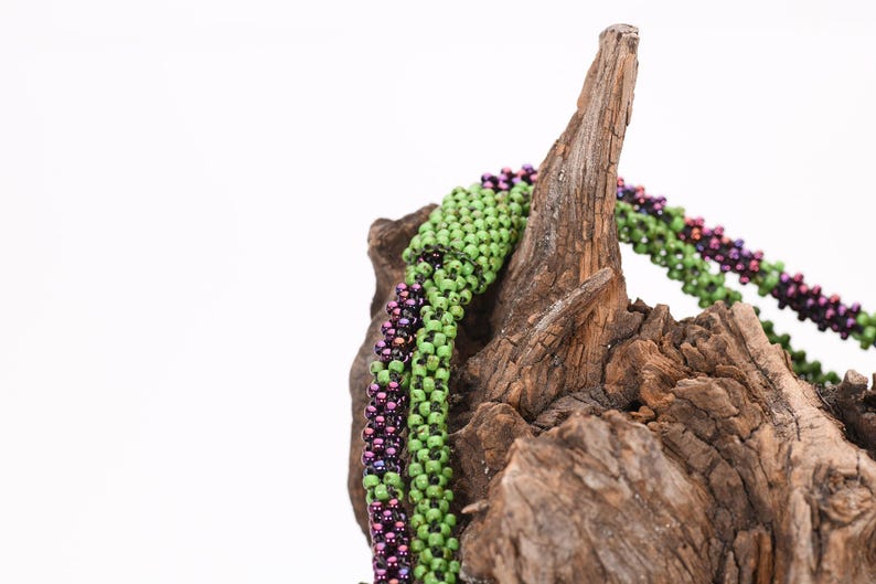 K&ouml;nnte beinhalten: Nahaufnahme einer gr&uuml;nen und violetten Perlenkette, die &uuml;ber einem St&uuml;ck Treibholz drapiert ist. Die Kette besteht aus kleinen, runden Perlen, die in einem komplizierten Muster miteinander verwoben sind.