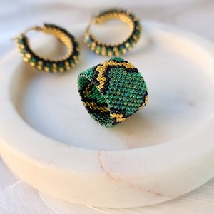 May include: A green and gold beaded ring with a geometric pattern. The ring is on a white marble surface. There are two matching earrings in the background.