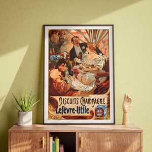 May include: Framed vintage advertisement for Biscuits Champagne Lefèvre-Utile. The artwork depicts a social gathering with people in elegant attire, enjoying drinks. The poster has a warm color palette with text in a decorative font.