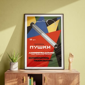 Op de afbeelding: Ingelijste vintage poster met een gedurfd geometrisch ontwerp met een grote, gestileerde sigaret. De poster heeft een kleurenschema van rood, blauw, geel en zwart. De tekst in het Russisch luidt "Pушки" en "25 stuks."