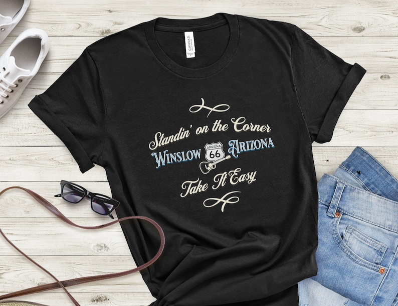 May include: Black t-shirt with a white graphic that says "Standin' on the Corner Winslow Arizona" with a Route 66 sign and a guitar. The text "Take It Easy" is below the graphic.