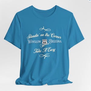 May include: A turquoise blue t-shirt with a white graphic design that says "Standin' on the Corner Winslow Arizona" with a Route 66 sign and a guitar. The text "Take It Easy" is below the graphic.