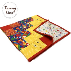 Puede incluir: Una colorida alfombra de juego para bebés con un borde rojo. La alfombra de juego está hecha de cuadrados de tela con diferentes diseños, incluyendo lunares, formas geométricas y abejas. La parte posterior de la alfombra de juego presenta un fondo blanco con letras coloridas del alfabeto.