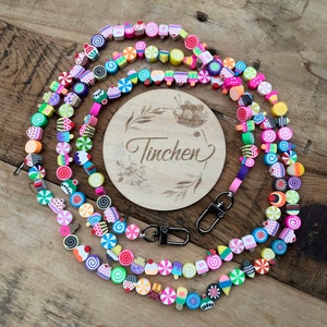 Könnte beinhalten: Ein farbenfrohes Schlüsselband mit Süßigkeiten-förmigen Perlen in Rosa, Grün, Gelb und Blau. Das Schlüsselband enthält eine Holzscheibe mit dem Namen "Tinchen" und zwei silberfarbene Verschlüsse zum Befestigen von Schlüsseln oder einem Ausweis.