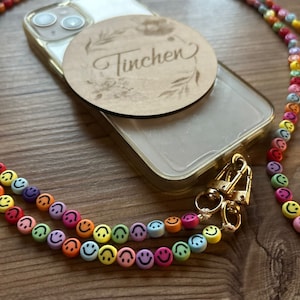Könnte beinhalten: Eine transparente Handyhülle mit einem goldenen Handyband. Das Band besteht aus bunten Smiley-Perlen. Ein Holz-Kreis mit dem eingravierten Wort "Tinchen" ist an der Rückseite der Handyhülle befestigt.